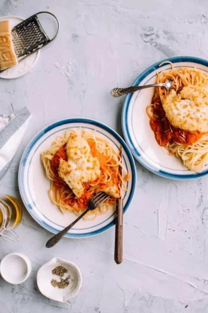 Baked chicken parmesan on top of spaghetti with tomato sauce on two plates with cutlery.