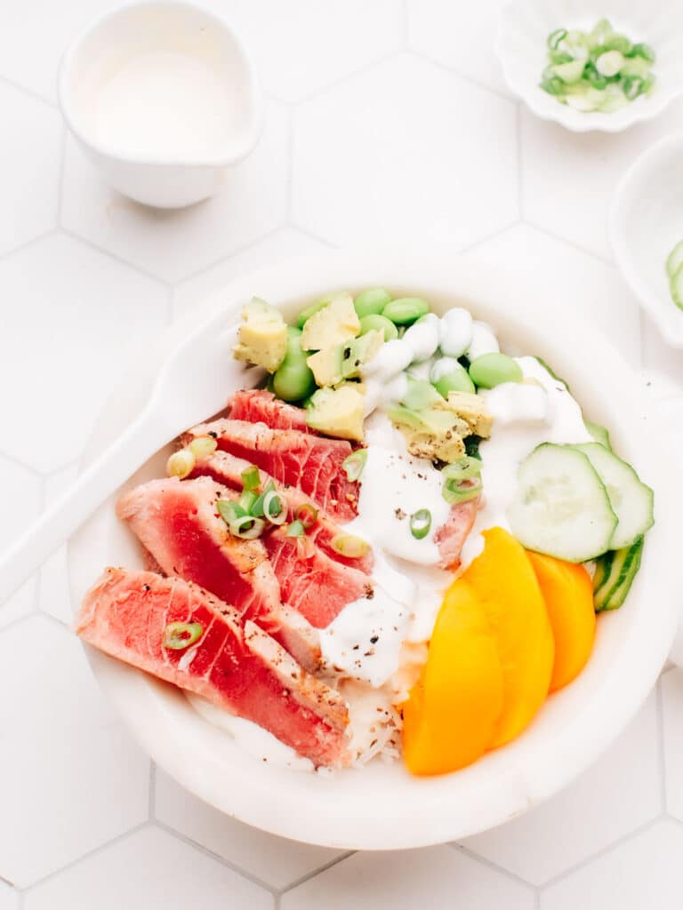 A delicious air-fried tuna steak is served over a bed of rice, topped with dressing, cucumber, edamame beans, and sliced mango. A white spoon rests on the bowl, and small bowls of spring onions are placed nearby.