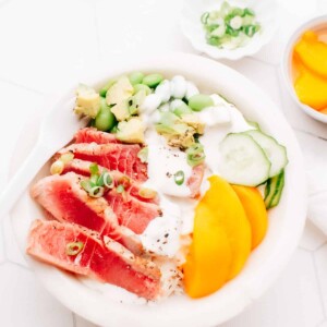 Sliced seared tuna in a poke bowl with peach slices, edamame, cucumber and green onions, drizzled with sauce.