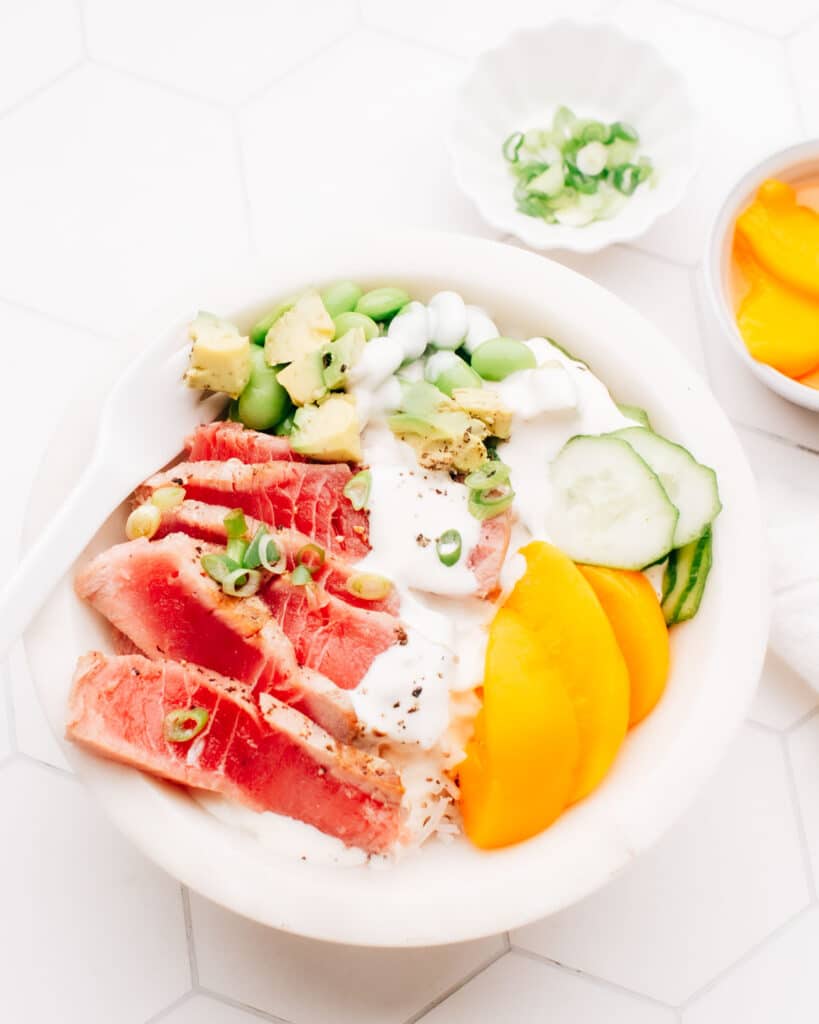 Sliced seared air fryer tuna in a poke bowl with peach slices, edamame, cucumber and green onions, drizzled with sauce.