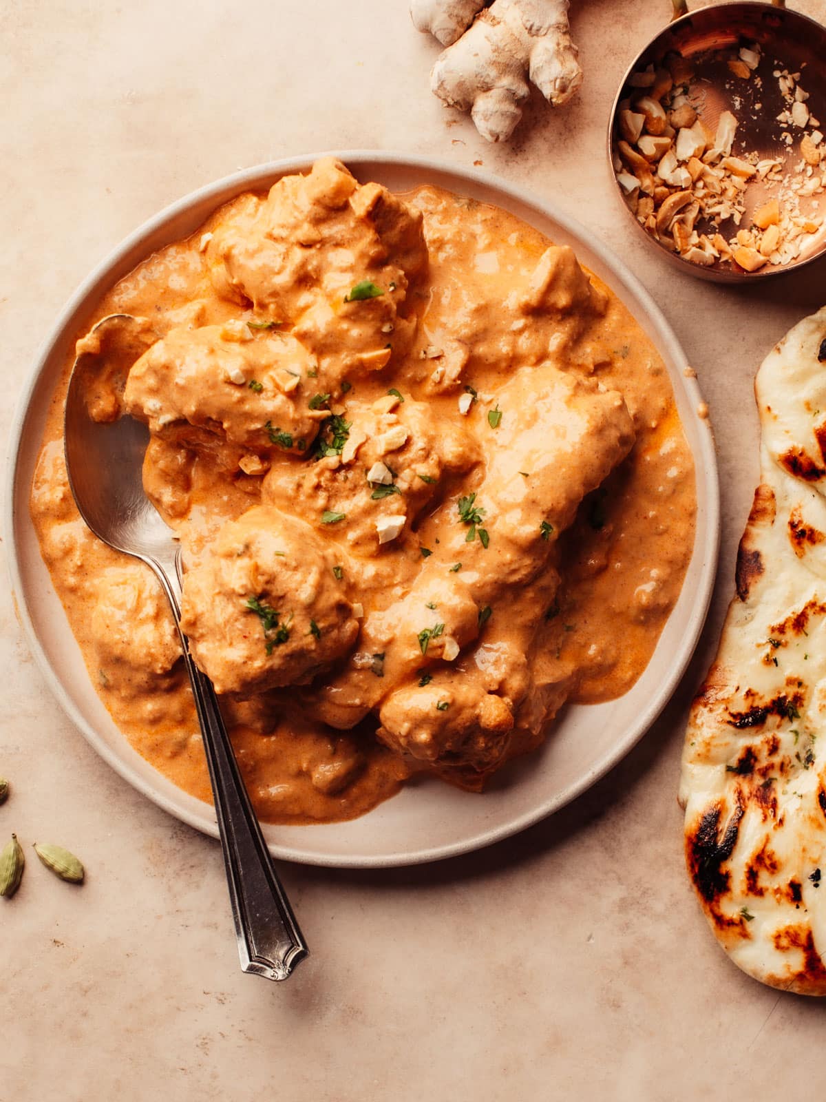 An aromatic and creamy chicken korma dish is beautifully presented in a large plate, garnished with fresh cilantro and nuts. A freshly baked homemade butter naan rests beside it, ready to be torn and dipped into the flavorful curry.