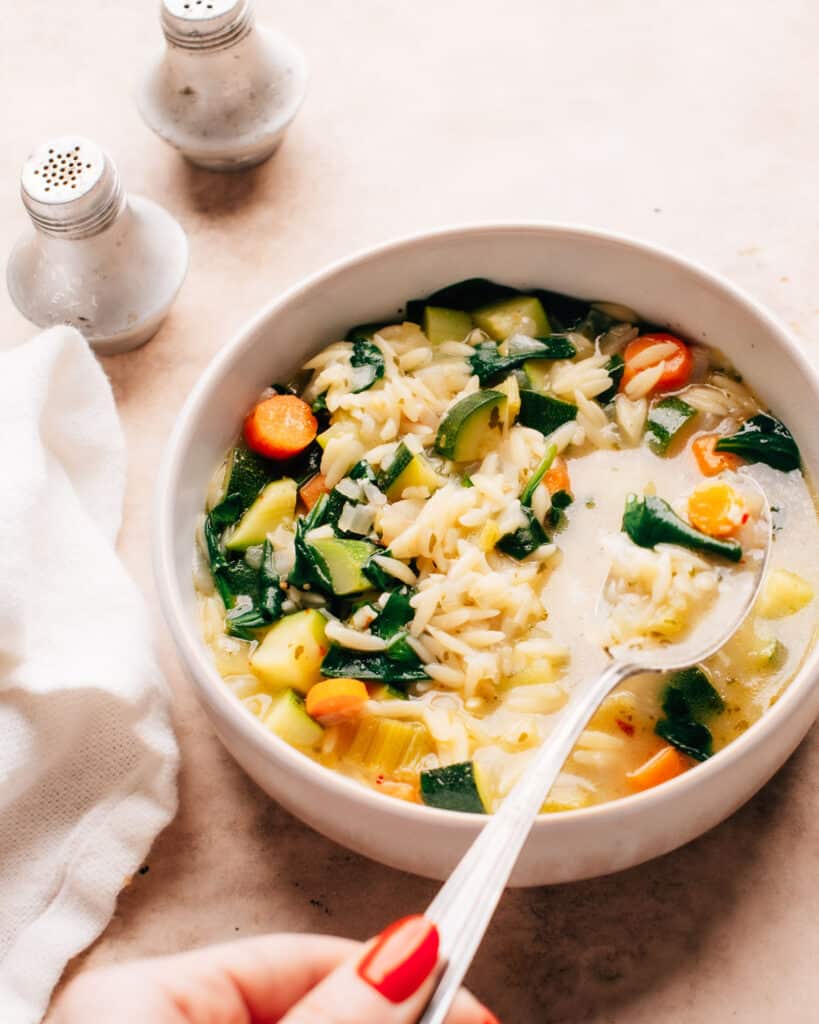 A white bowl filled with steamy vegetable orzo soup, spoon ready and waiting to be devoured