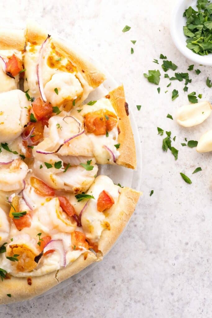 Half view of delicious seafood pizza on a kitchen countertop, topped with shrimp, scallops, white sauce and a sprinkle of parsley