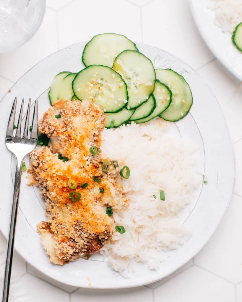 A golden panko-crusted cod fillet rests atop a bed of fluffy white rice and sliced cucumbers, served on a white plate.
