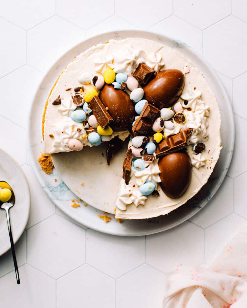 A delicious No-Bake Easter Egg Cheesecake presented on a large plate. The cheesecake is beautifully decorated with colorful Easter eggs, adding a festive touch to the dessert. A single slice of cheesecake has been cut out, revealing the smooth and creamy texture of the dessert.