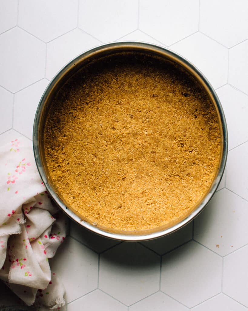 The bottom of a springform pan, with a crumbly crust made from graham crackers, is pressed firmly into the bottom. The crust is evenly distributed and tightly packed, providing a solid base for a delicious no-bake easter egg cheesecake recipe.