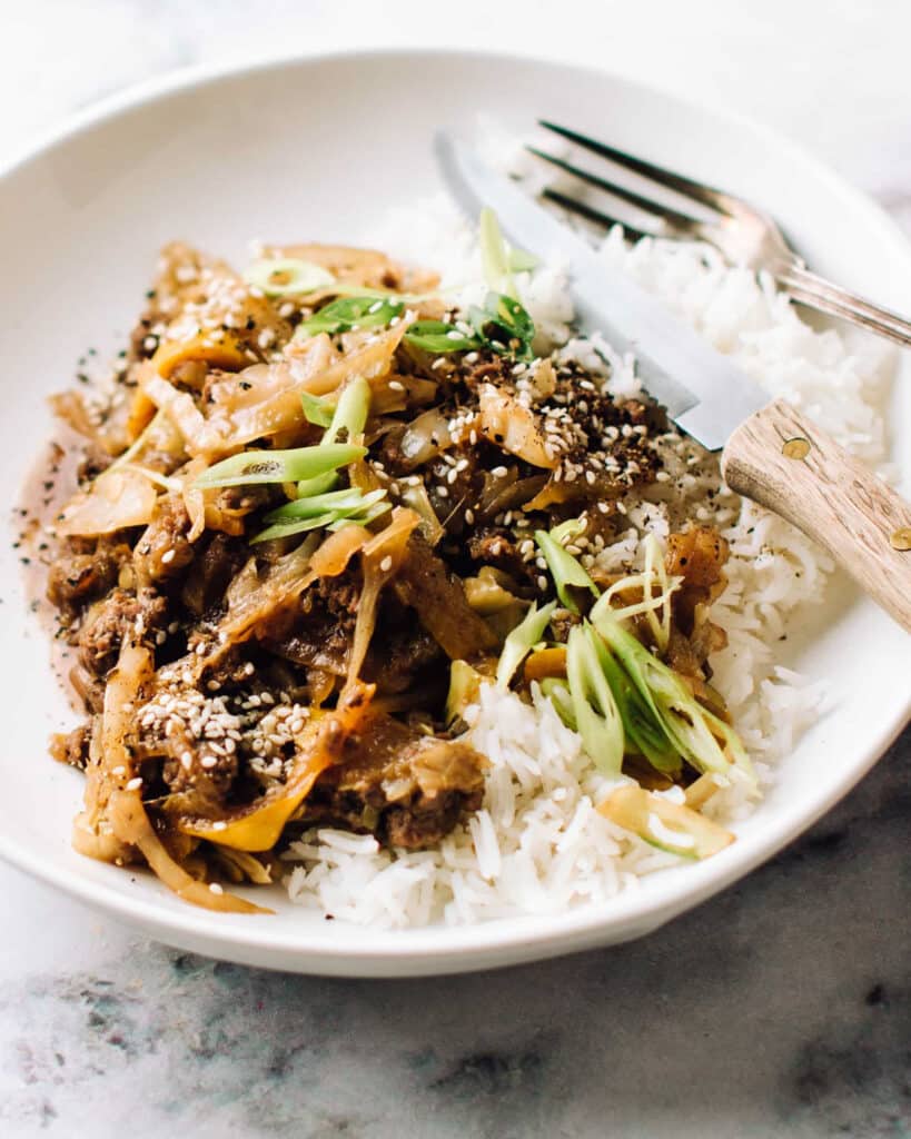 A white plate filled with a delicious ground beef stir fry, served over a bed of fluffy rice. The stir fry includes tender pieces of ground beef, sautéed with vegetables such as onions, zucchini and green cabbage.