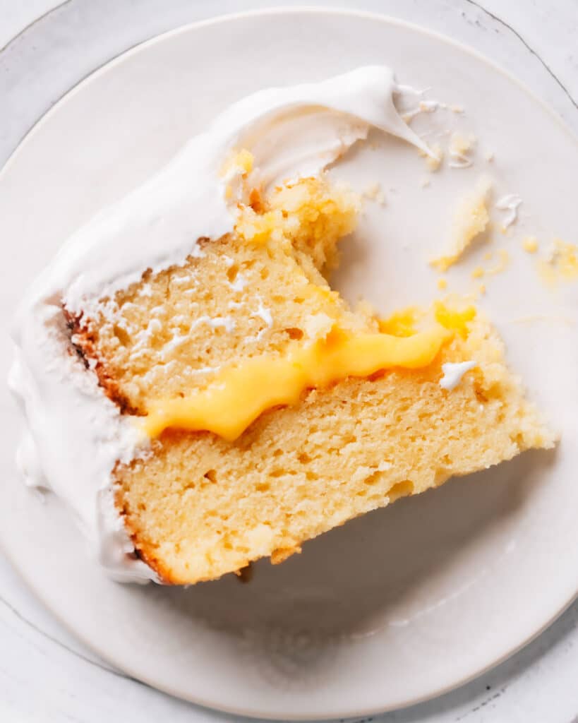 Slice of lemon curd cake on white plate with layers of lemon cake, tangy lemon curd filling, and fluffy white frosting. A portion cut with a fork is visible.