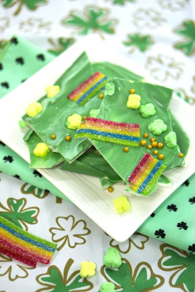 Stacked St. Patrick's Day leprechaun bark on a white plate. The bark is made of layers of chocolate and studded with festive sprinkles, gold dust, and shamrock-shaped candies. 