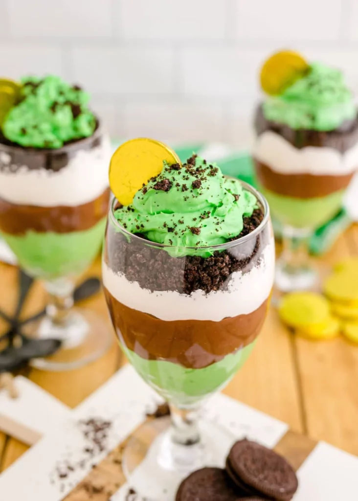 St. Patrick's Day Pudding Parfait, presented in a tall glass with a chocolate gold coin on top. The dessert consists of layers of creamy vanilla pudding and vibrant green pudding, creating a festive color scheme.