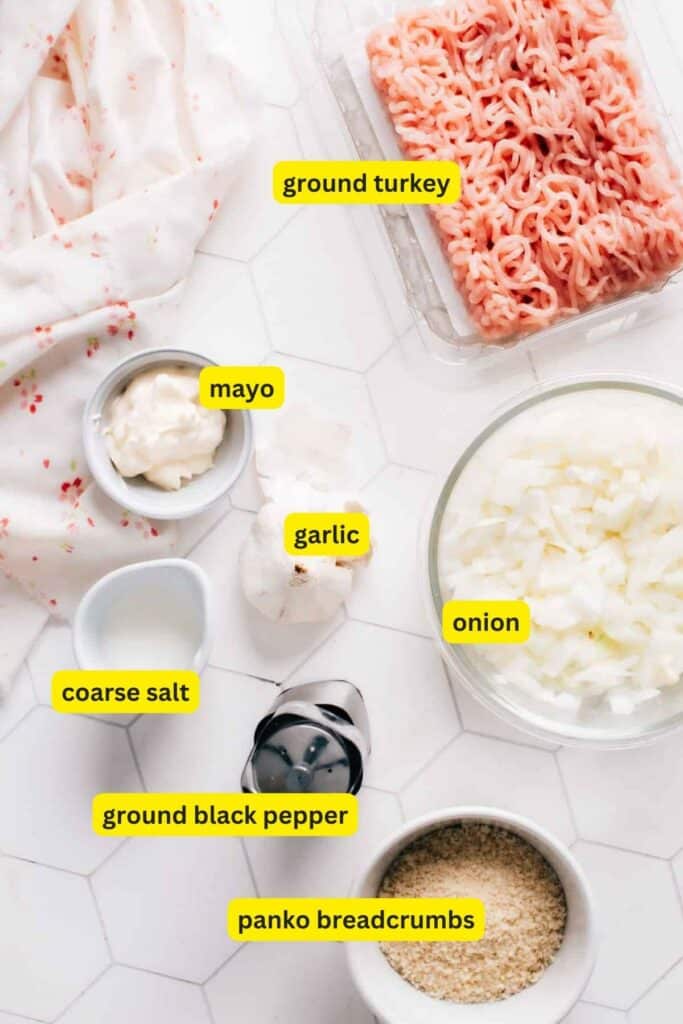 Ingredients for turkey burgers laid out on a white kitchen countertop, including ground turkey, slices onions, mayo, salt, pepper and panko breadcrumbs.