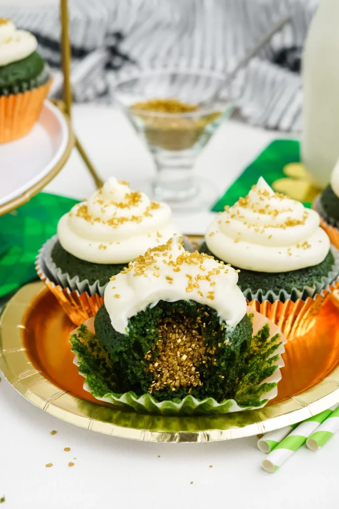 Three green velvet cupcakes with cream cheese frosting, placed on a golden plate. One of the cupcakes is sliced open, revealing a moist, green crumb with gold sparkles inside. 