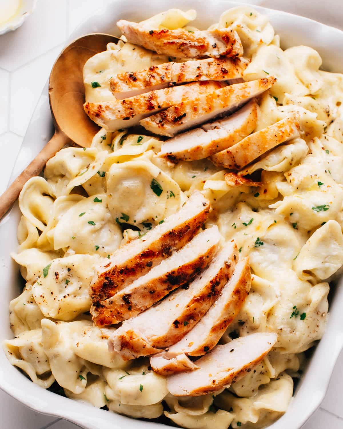 Asiago tortellini Alfredo with grilled chicken served in a white baking dish on a wooden table. The dish is garnished with minced chives and parsley.