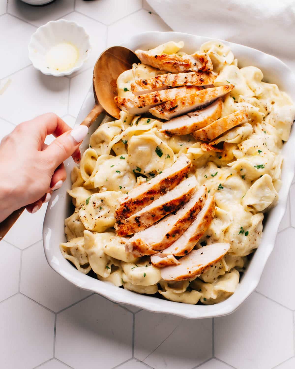 Asiago tortellini Alfredo with grilled chicken served in a white baking dish on a wooden table. The dish is garnished with minced chives and parsley