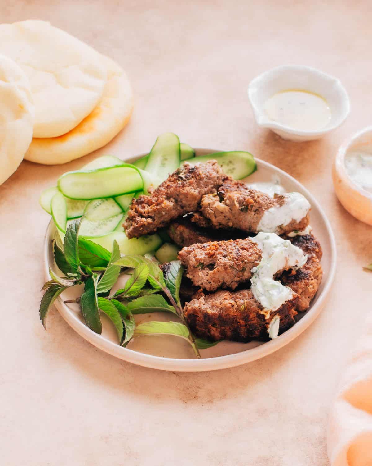 A plate of beef kafta kabobs with mint and sliced cucumber. Turkish bread is placed behind the plate.
