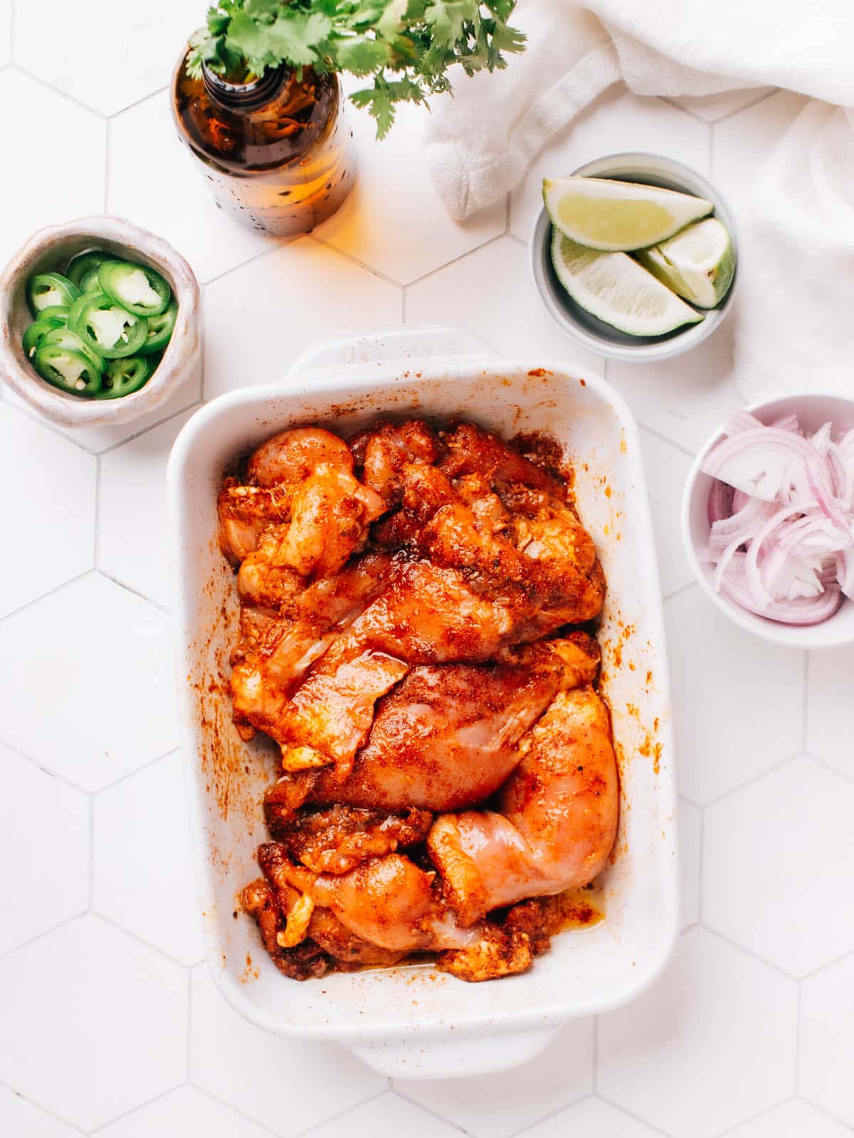 The marinated chicken thighs are left to rest at room temperature for 30 minutes. There are individual bowls of sliced jalapenos, onions, and limes beside the chicken bowl.