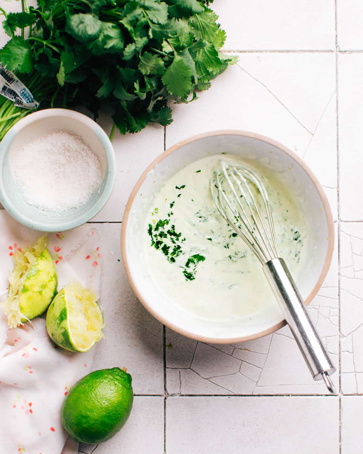 Freshly made cilantro lime crema in a small bowl with a whisk. Limes, kosher salt, and cilantro are placed nearby.