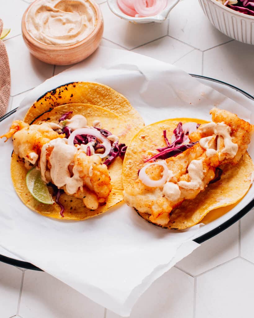 Two delicious Baja fish tacos artfully presented on parchment paper, drizzled with flavorful Baja sauce and accompanied by a bed of crispy cabbage slaw and pickled onions.