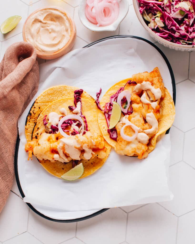 Two delicious Baja fish tacos artfully presented on parchment paper, drizzled with flavorful Baja sauce and accompanied by a bed of crispy cabbage slaw.