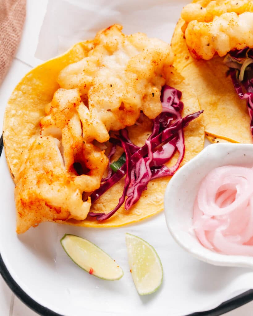 Fish tacos with a flavorful cabbage slaw are presented, accompanied by a small cup of sliced onions and lemon wedges for that extra zesty touch.