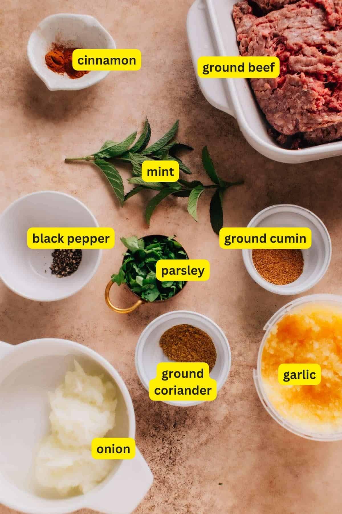 Ingredients for Beef Kafta Kabob arranged on a kitchen countertop: ground beef, cinnamon, mint, black pepper, parsley, ground coriander, ground cumin, garlic, and onion.