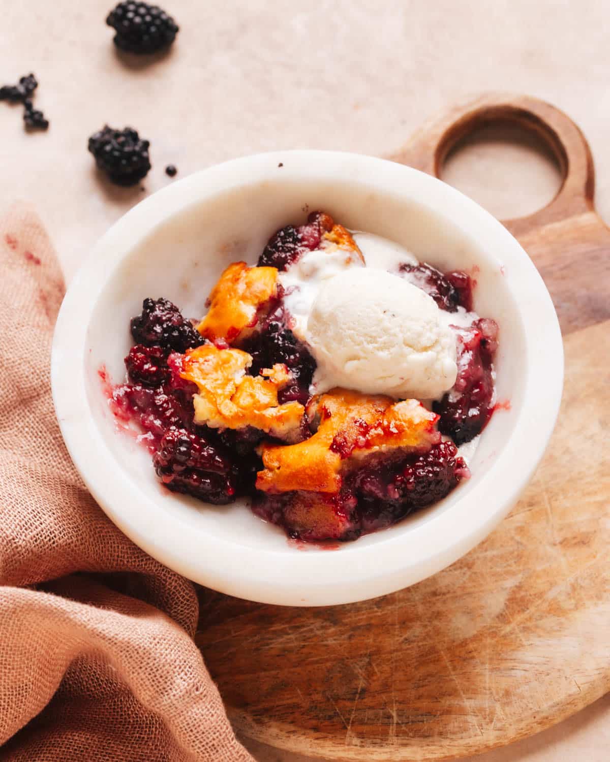 Fresh blackberry cobbler topped with a dollop of vanilla ice cream in a white dish. The dish is set on a wooden cutting board with a kitchen cloth. Fresh blackberries are displayed in the background.