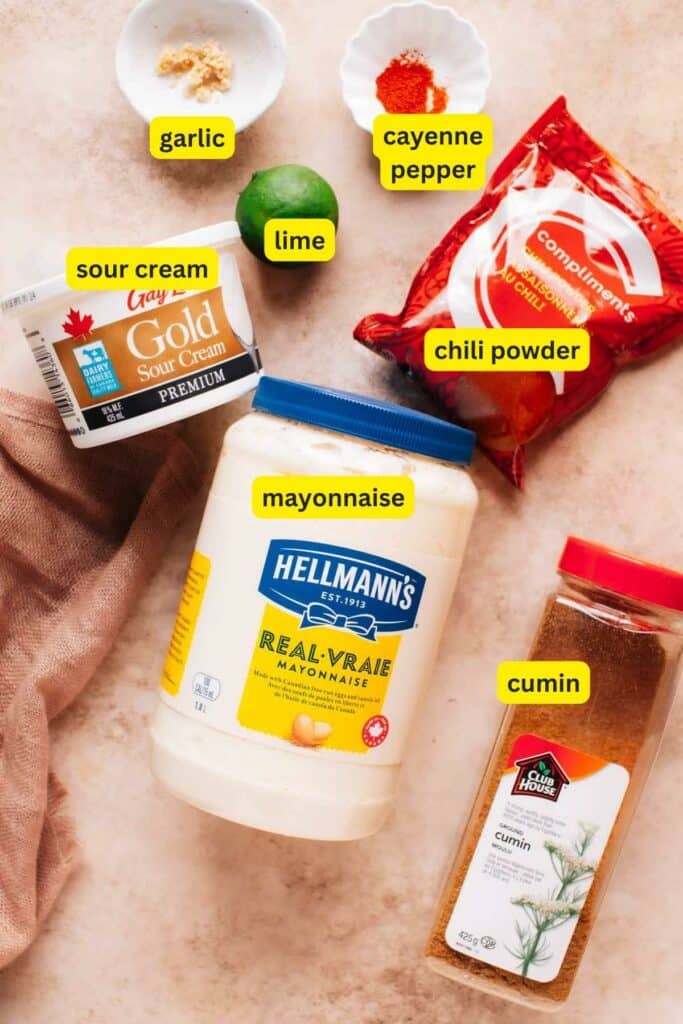 Ingredients for Baja sauce are laid out on the kitchen countertop, including mayonnaise, garlic, sour cream, lime, chili powder, cumin, and cayenne pepper.