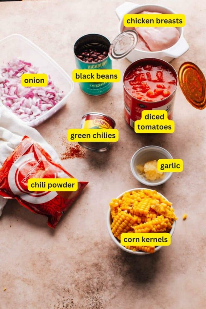 The ingredients for cream cheese chicken chili on a kitchen countertop include chili powder, chicken breasts, onion, garlic, black beans, corn kernels, green chilies, and diced tomatoes.