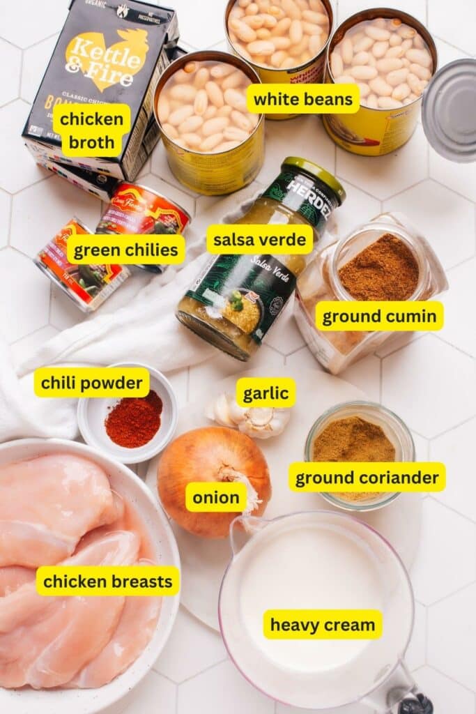 The ingredients for white chicken chili on a table include chili powder, chicken breasts, onion, garlic, white beans, green chilies, chicken broth, salsa verde, heavy cream, ground cumin, and ground coriander.