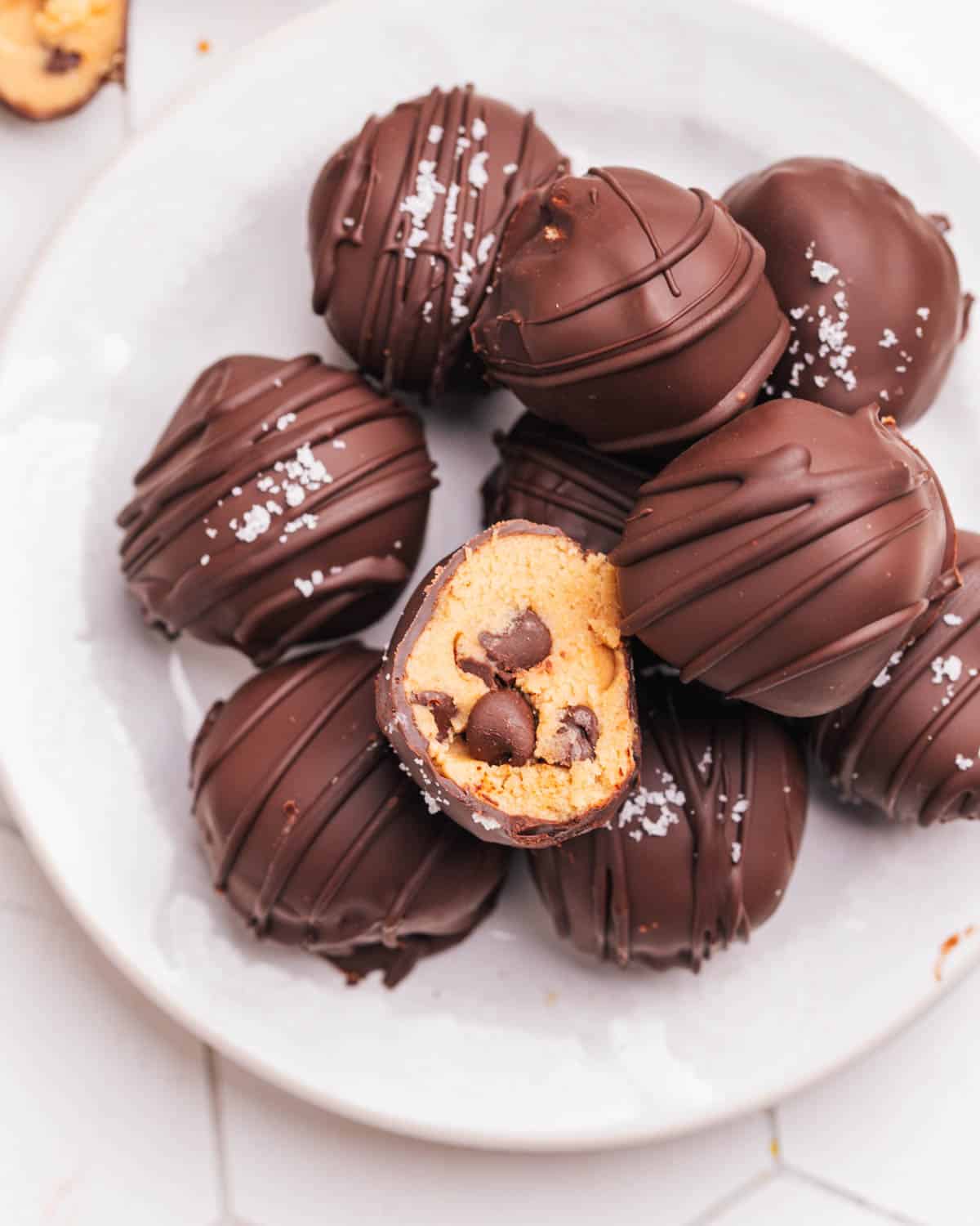 Cookie dough bites covered in chocolate in a pile on a plate. The top one has a bite taken.