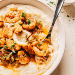 A bowl of shrimp and grits with seasoned shrimp, a bacon pan sauce, and a lemon slice over creamy cheesy grits.
