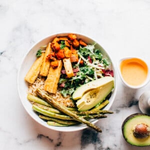 Rice bowl with asparagus, avocado, sweet potato, tofu and greens with sauce and more avocado to the side.