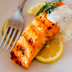 Dill sauce spooned over a filet of grilled salmon on a plate with lemon slices.