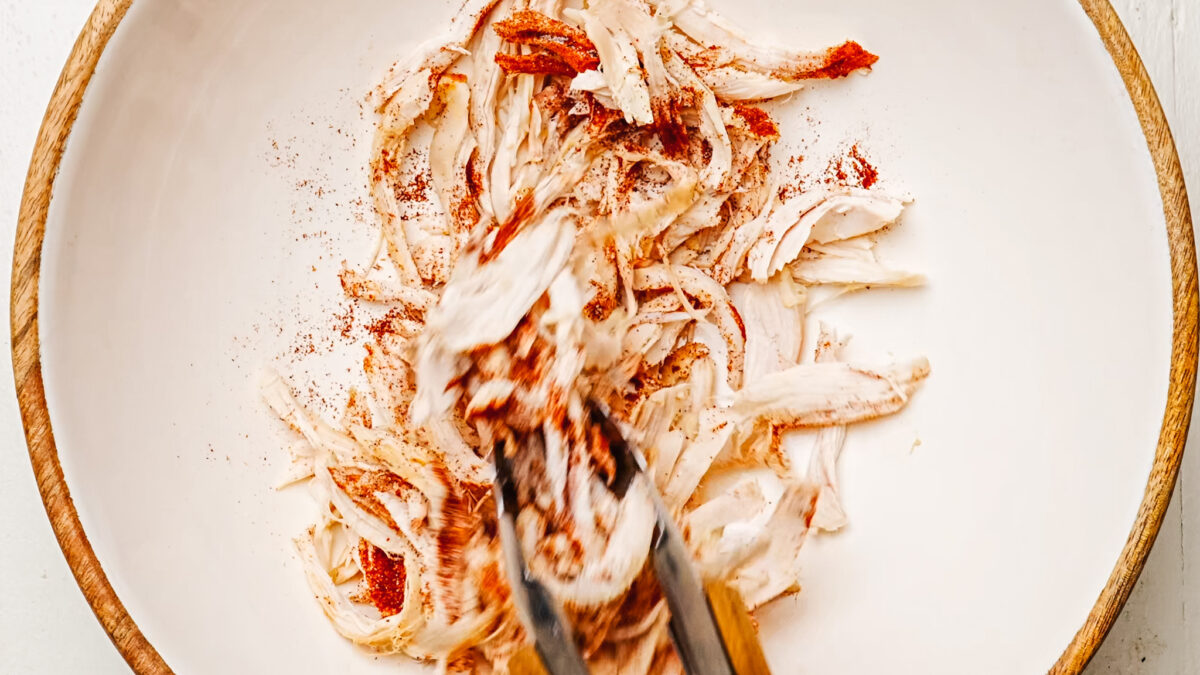 Shredded chicken being tossed with chili powder in a bowl.
