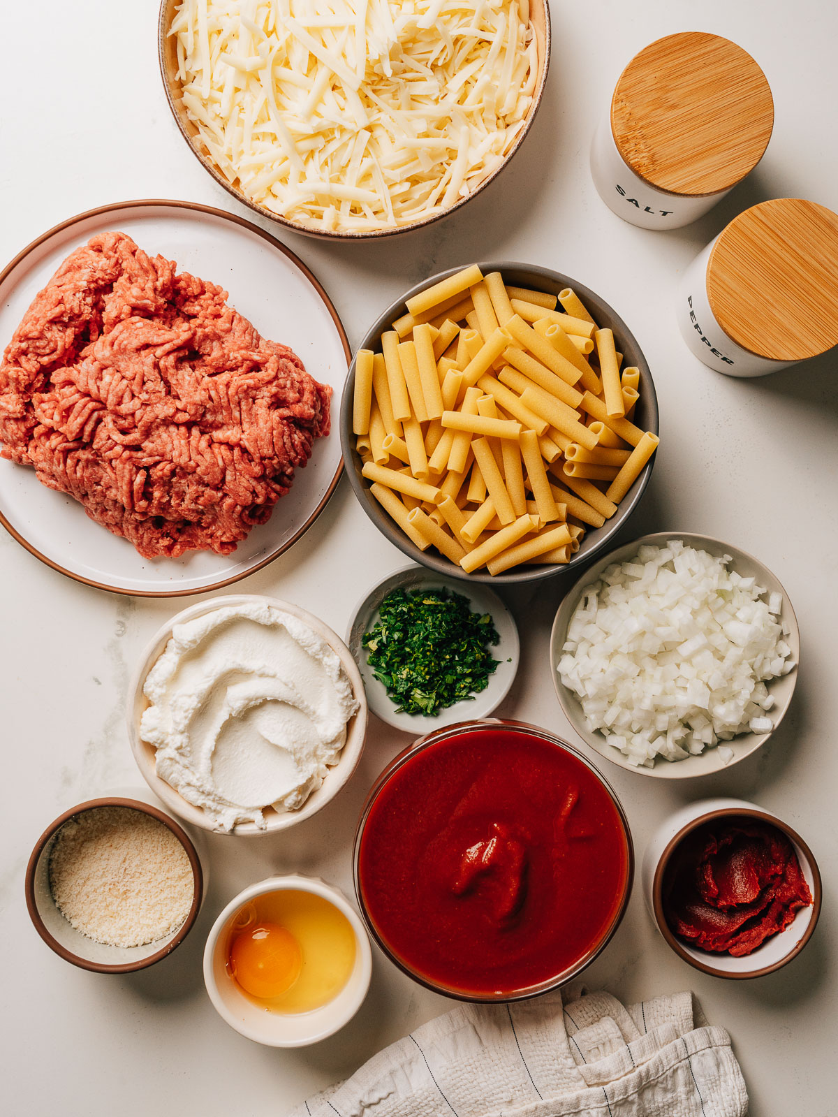 Ingredients for baked ziti including ziti pasta, ground beef, ricotta, mozzarella, tomatoes, and fresh herbs