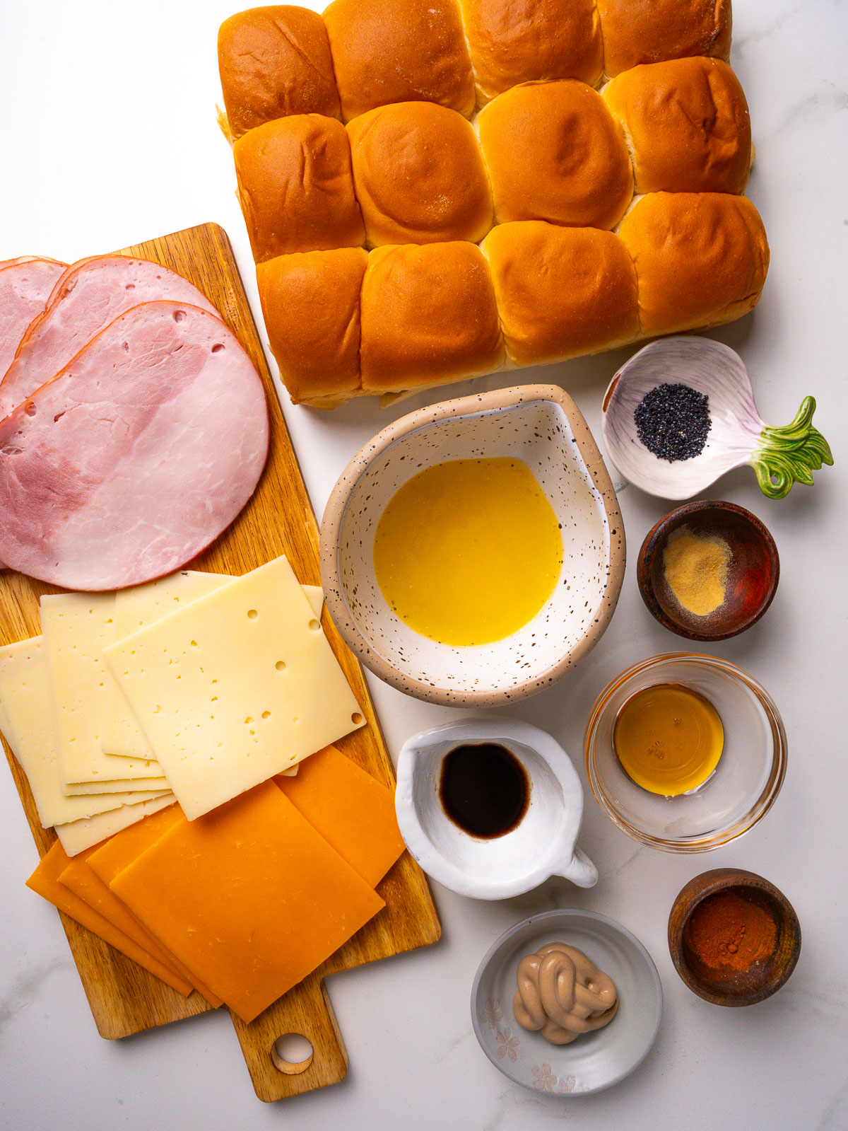 Overhead view of ingredients for Hawaiian ham and cheese sliders: sweet Hawaiian rolls, deli ham, Swiss and cheddar, butter, Dijon, Worcestershire and poppy seeds.