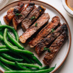 Sliced garlic butter steak on a plate with green beans and a dish of garlic butter.