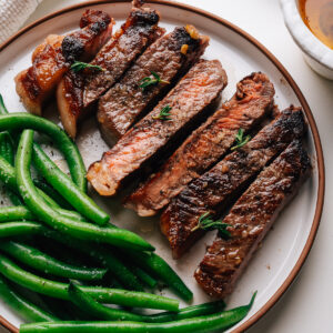 Sliced garlic butter steak on a plate with green beans and a dish of garlic butter.