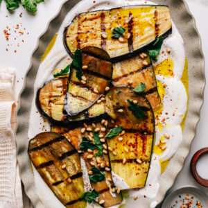 Grilled eggplant strips arranged over whipped feta with mint and pine nuts on a platter.