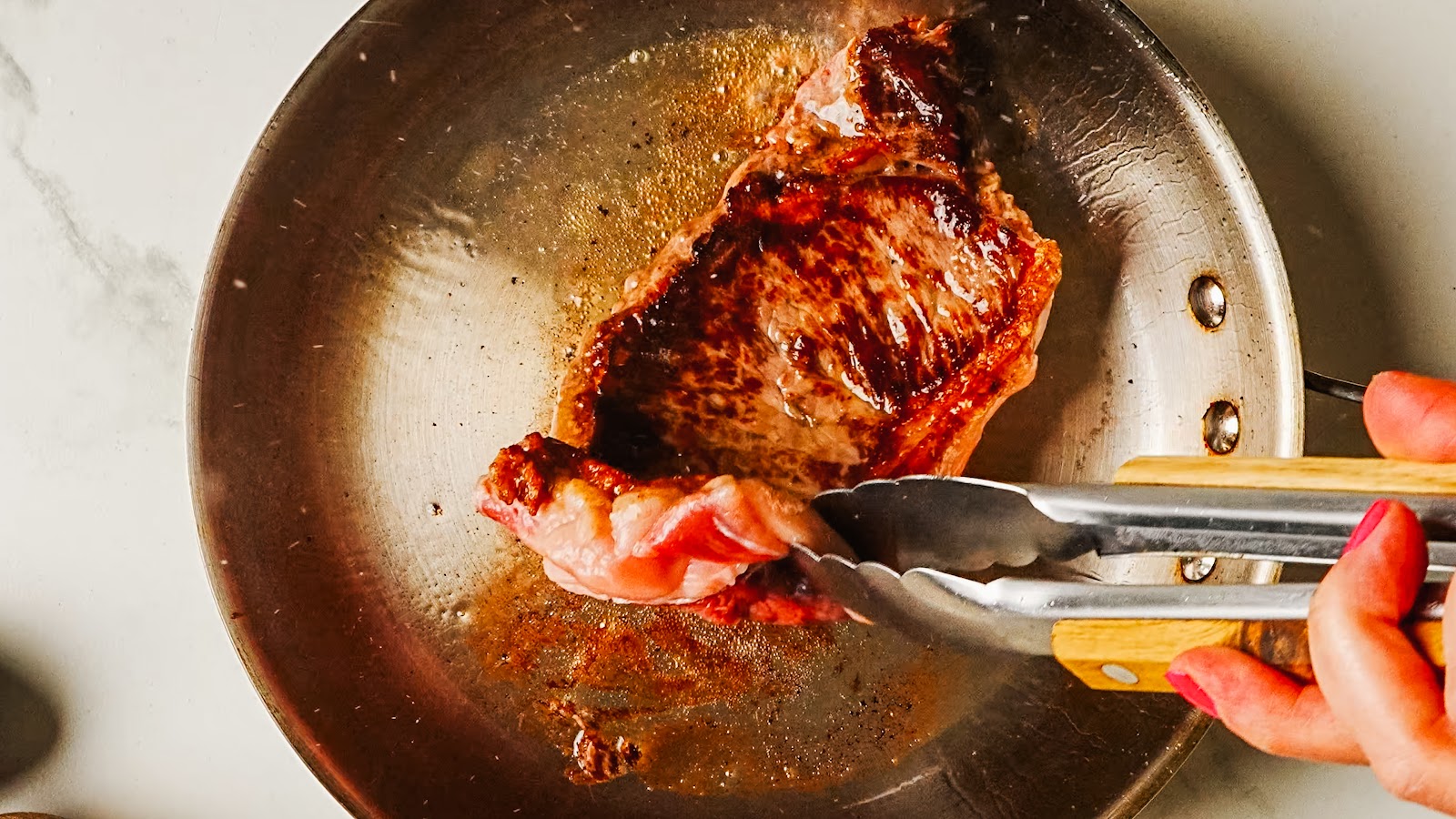 Steak searing with a deep brown crust, lifted with tongs.