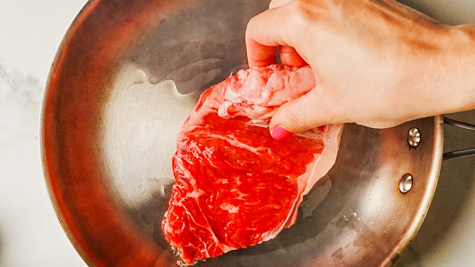 Seasoned steak laid into a hot stainless pan to sear.