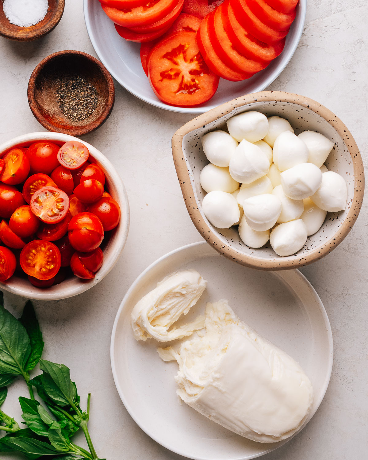 Caprese Salad recipe ingredients with cherry tomatoes, mozzarella and basil on a white surface.