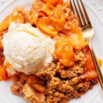 A serving of peach crumble on a white plate beside a scoop of vanilla ice cream.