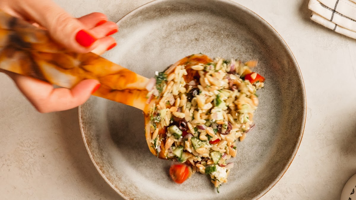 A wooden spoon lifting a serving of orzo pasta salad from the bowl.