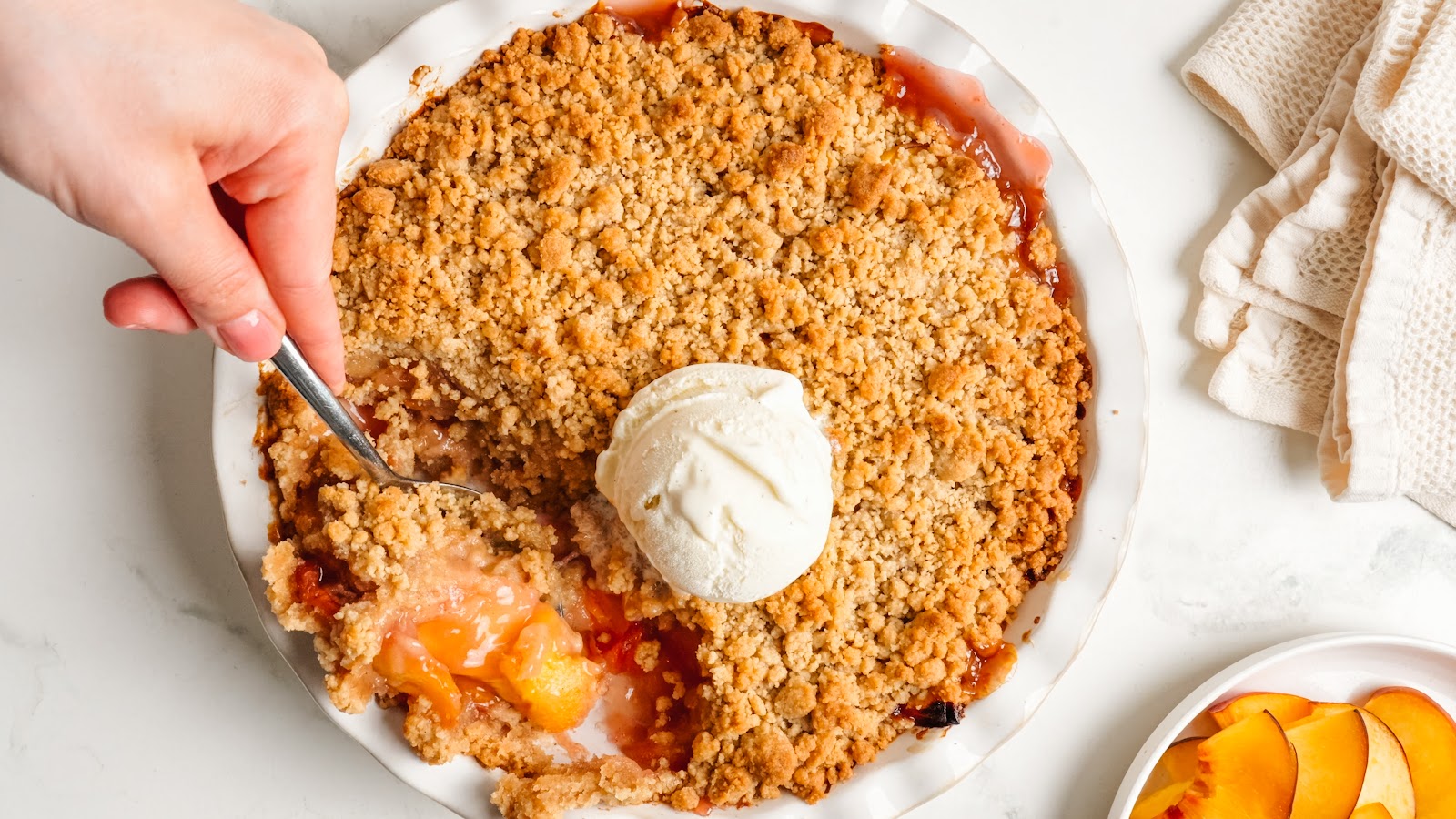 Baked peach crumble in a white pie dish with a scoop of vanilla ice cream melting on top.