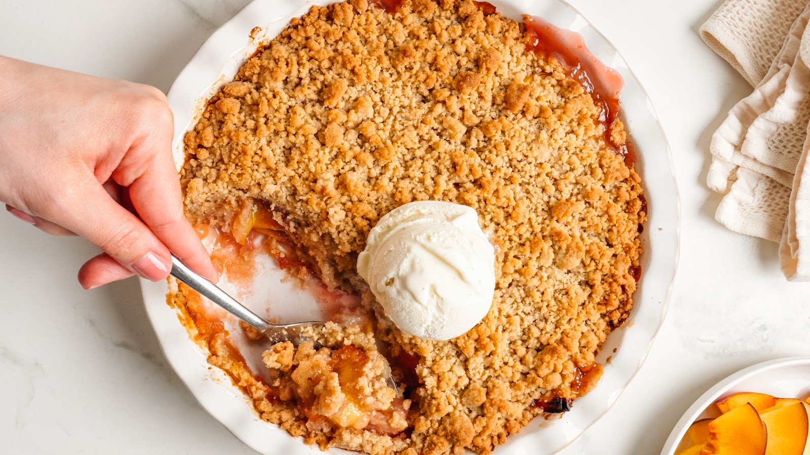 A spoon lifting golden brown oat crumb topping and jammy peaches from the dish.